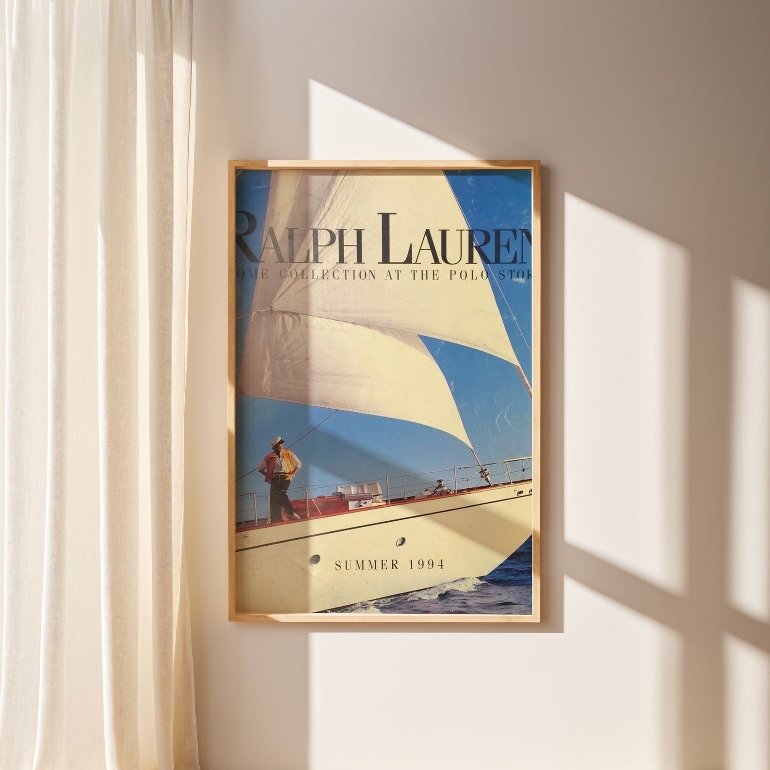 Vintage sailboat poster Ralph Lauren style Summer 1994 in wooden frame on beige wall with sunlight shadows.